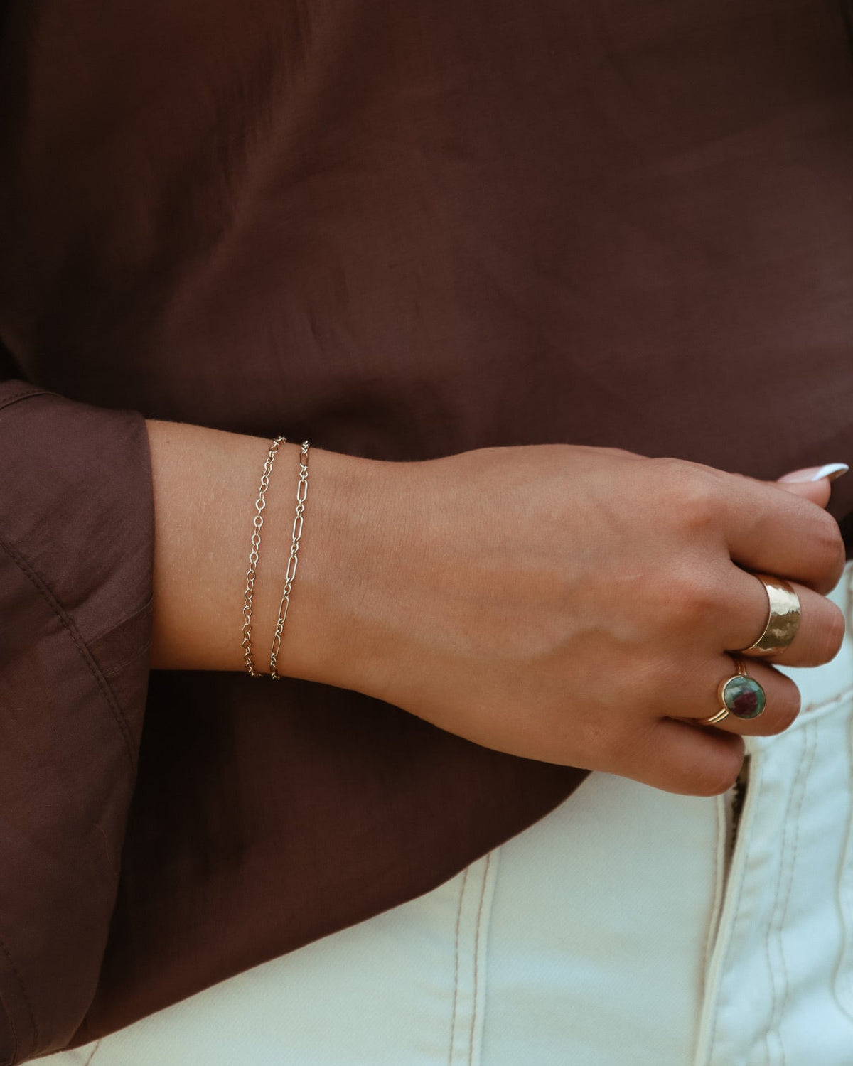 Close-up of layered gold chain bracelets and statement rings styled with a brown blouse and cream pants.