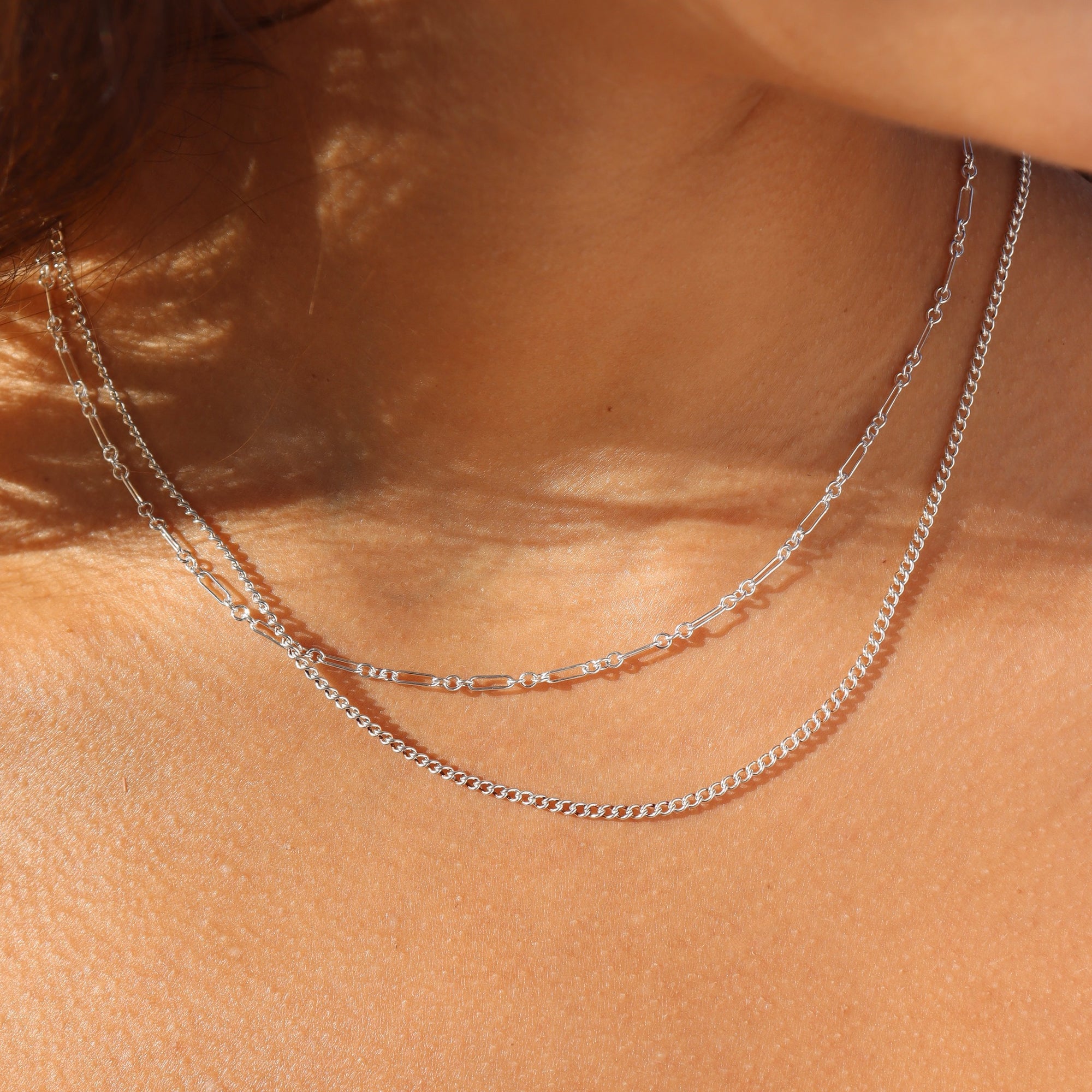 Extreme close-up shot of a woman's neckline highlighting two layered sterling silver chains in bright natural sunlight: a fine bar-and-link chain layered above a slightly longer delicate silver curb chain.