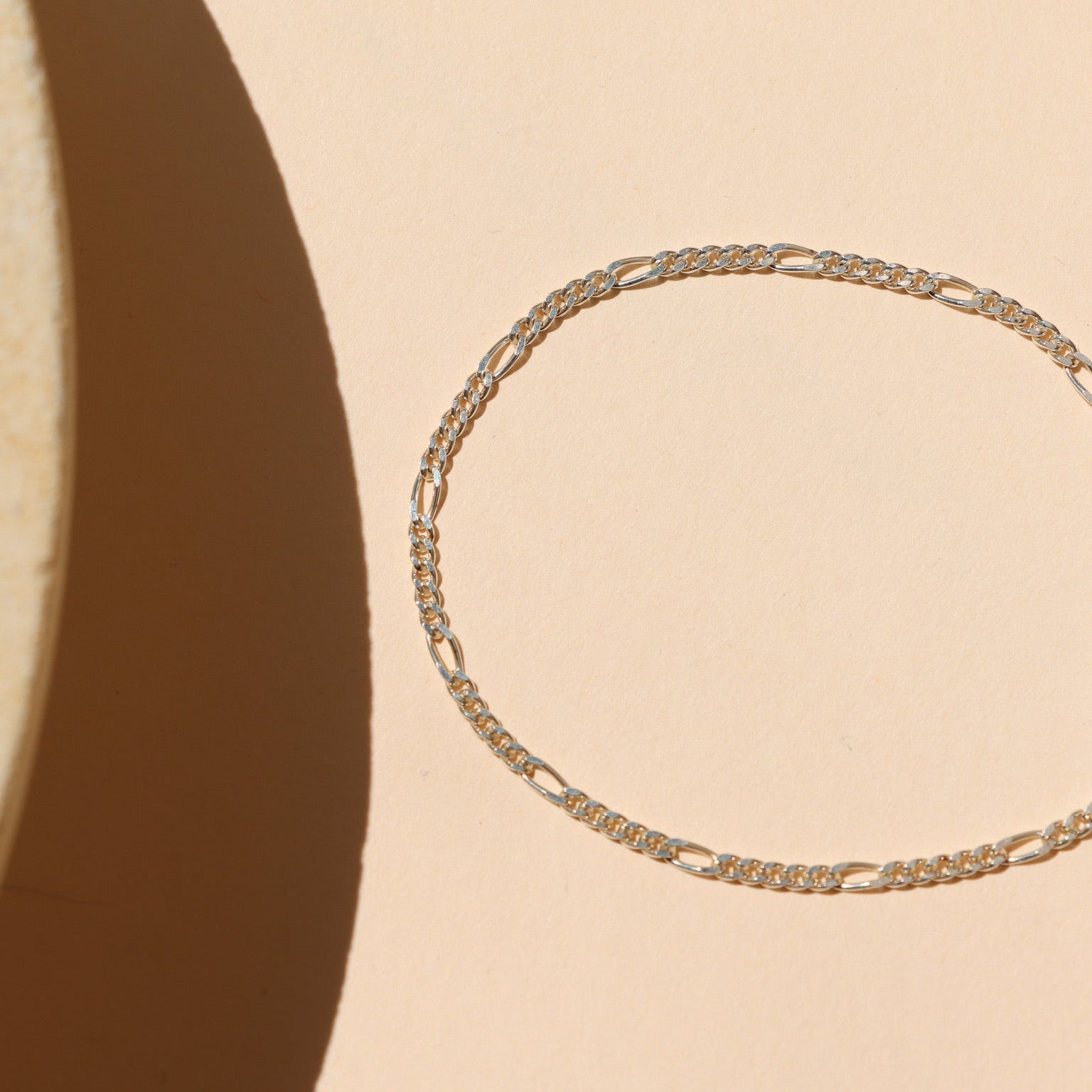 Minimalist sterling silver Figaro chain bracelet featuring alternating short round links and elongated oval links, laid flat on a peach-beige surface with geometric natural light shadows.