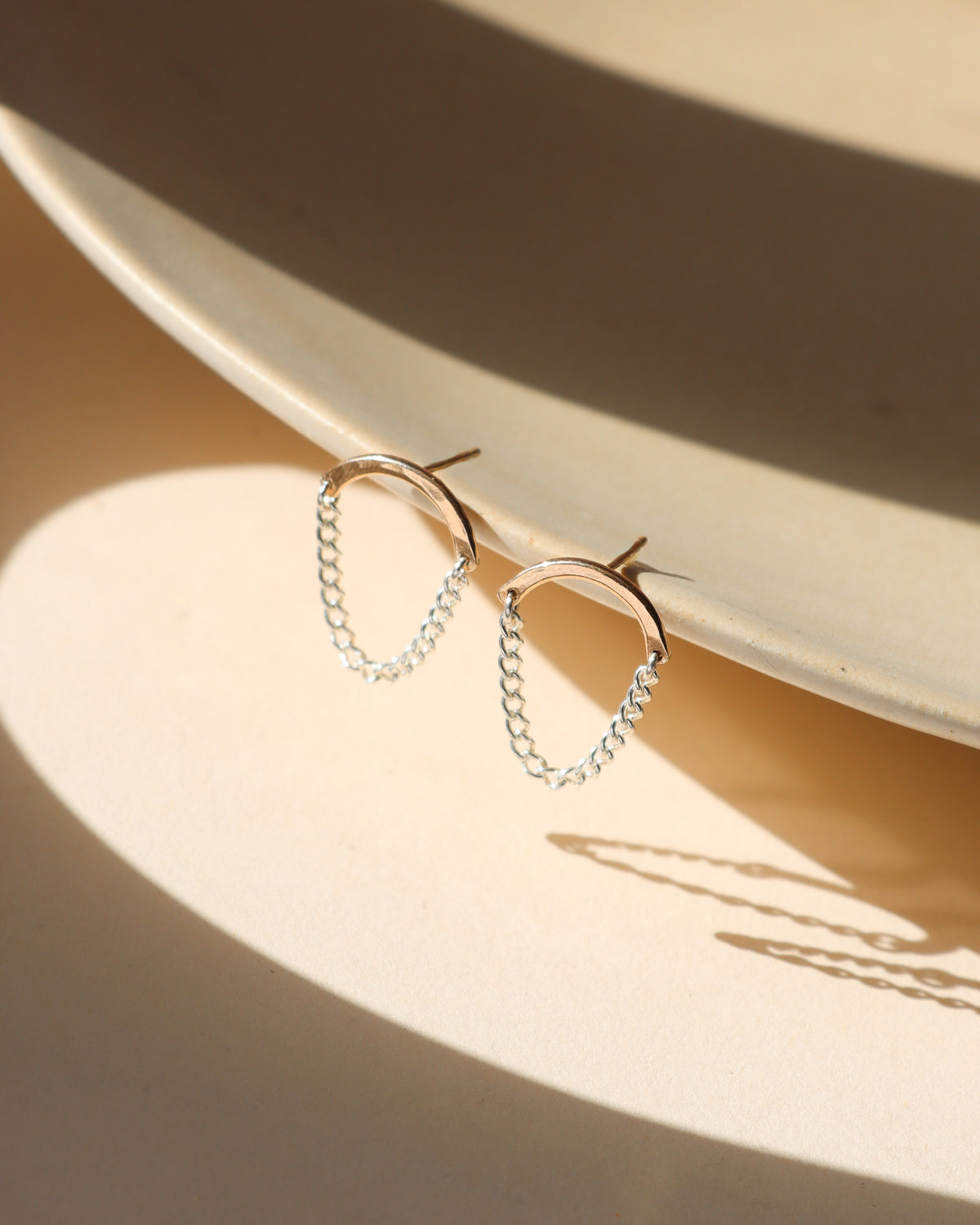 Pair of sterling silver chain drop earrings featuring curved silver bars connected by delicate draped chains, resting along the edge of a cream ceramic dish in soft sunlight.
