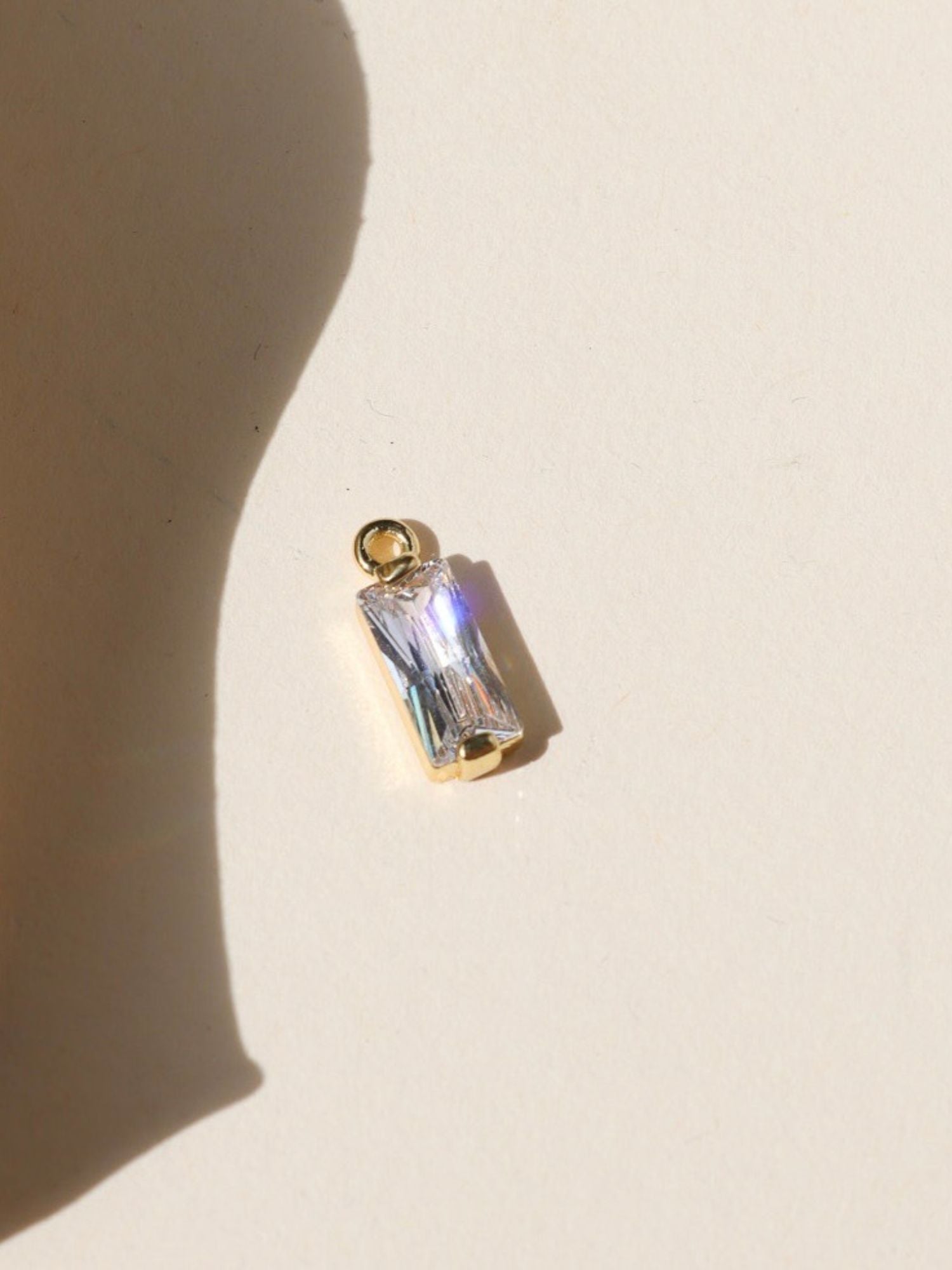 Gold charm featuring a clear baguette-cut crystal, photographed on a warm neutral surface with soft natural light and shadows.