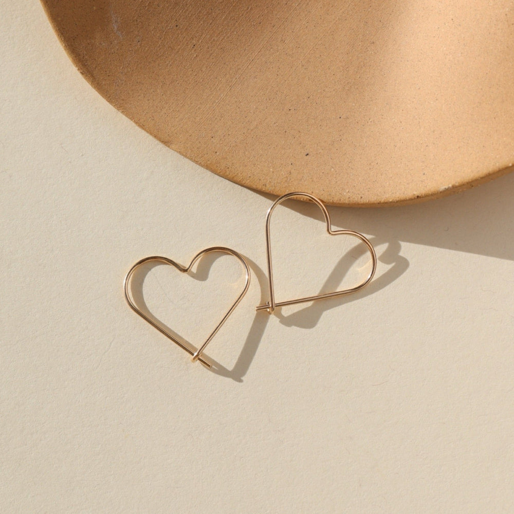 14k gold fill open heart slide earrings. This earring features a gold wire that is shaped into an open heart, with a hook closure. Earrings are displayed on a cream backdrop, rested on a tan dish. 