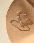 sterling silver open heart slide earrings. This earring features a silver wire that is shaped into an open heart, with a hook closure. Earrings are displayed on a cream backdrop, rested on a tan dish. 