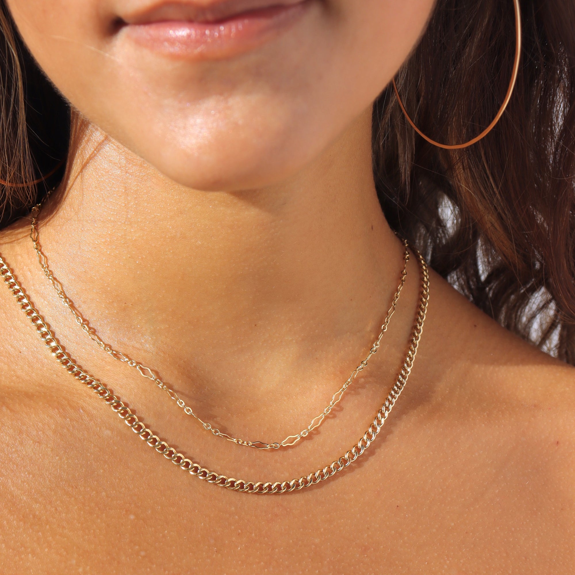 Close-up shot of a woman's neckline highlighting layered gold jewelry in warm sunlight: a delicate open-link chain styled above a classic gold curb link chain, paired with thin, large gold hoop earrings