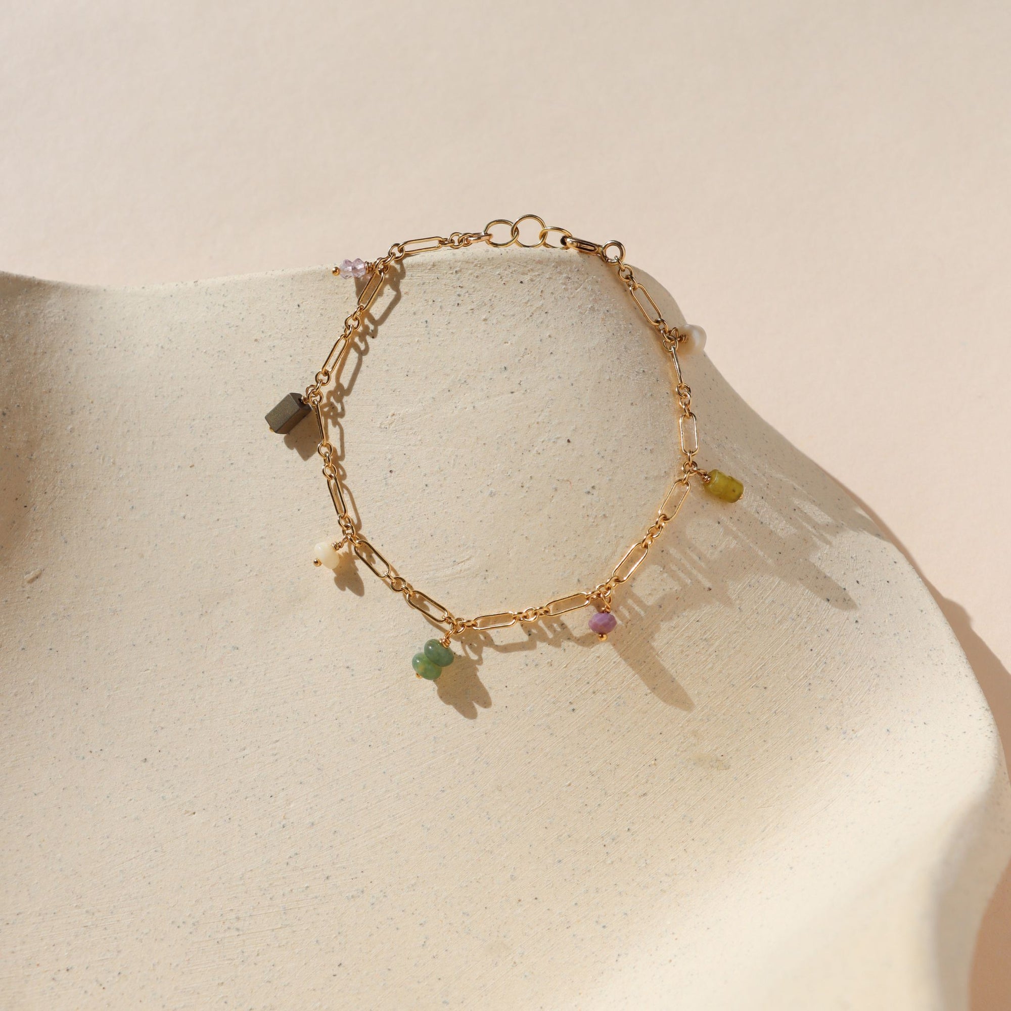 Gold chain linked bracelet with gemstone charms evenly spaced across it, laid on a beige dish in the sunlight