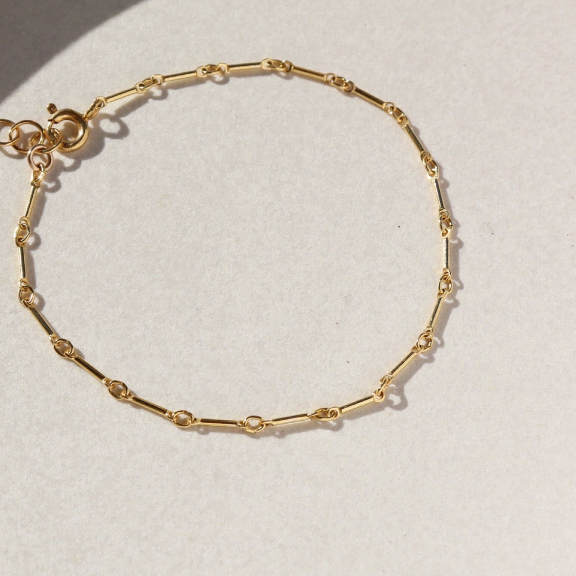 Close-up product photo of the Dot + Dash Bracelet, a delicate 14k gold fill chain bracelet with elongated bar links, photographed on a light stone surface in natural sunlight.