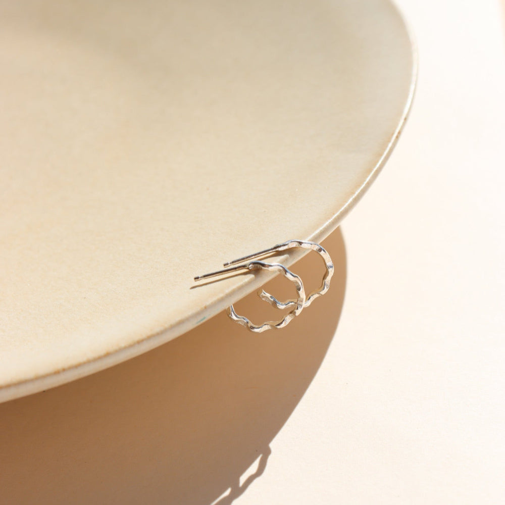 A wave like sterling silver textured huggie earring, displayed on a tan dish in the sunlight. 
