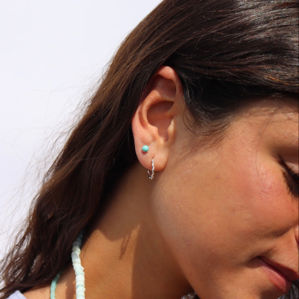 Model wearing a wave like sterling silver textured huggie earring. She pairs her hoop with a 4mm sterling silver turquoise stud. 