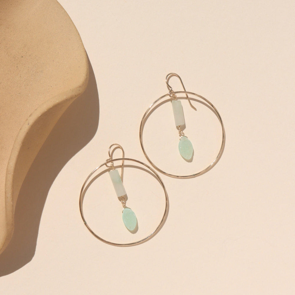 A 14k gold fill hoop with druzy + burma jade gemstones dropping from the ear post. The earrings are displayed on a cream back drop in the sunlight.