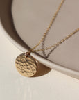 14k gold fill sun drop necklace laid on a tan plate in the sunlight. This necklace features our simple chain met by a hammered coin, making it look like the moon.