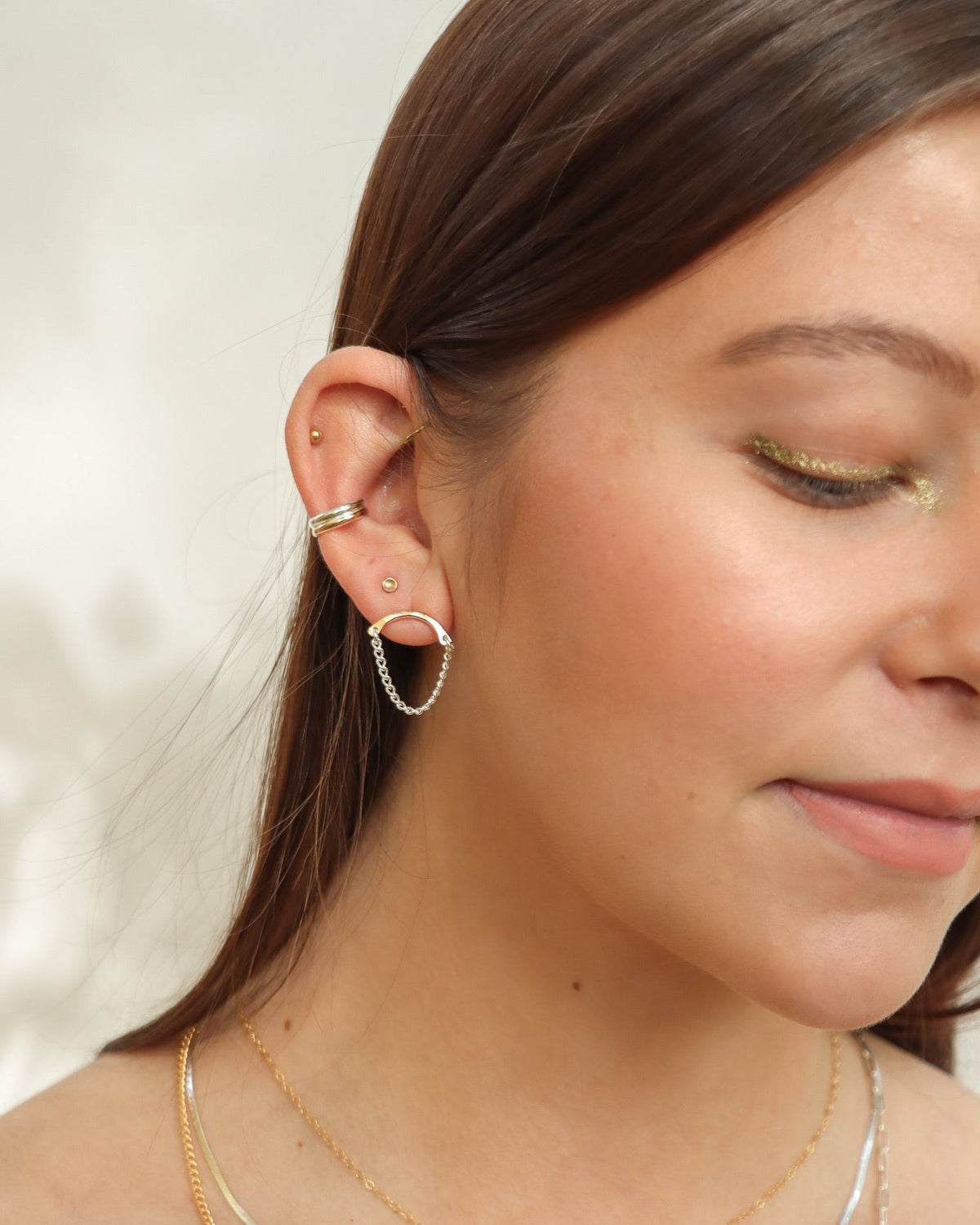 Close-up of a model wearing Token Jewelry's mixed metal Balance Studs, featuring a gold curved bar with a delicate silver chain drop, styled with layered gold and silver necklaces and mixed metal ear cuffs.