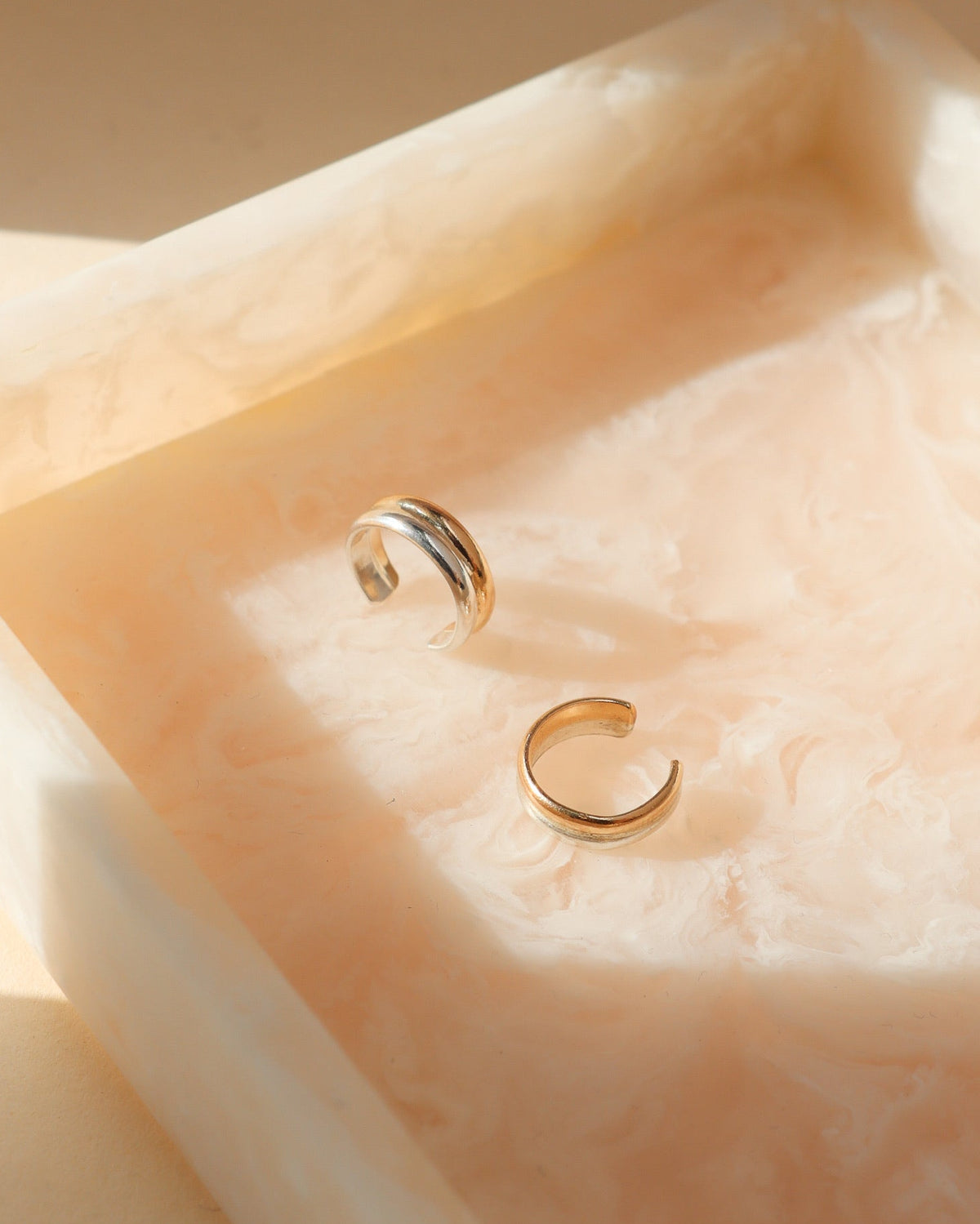 Two Token Jewelry ear cuffs in gold and mixed metal finishes, featuring smooth, layered bands and an open cuff design, displayed on a peach-toned marbled tray.