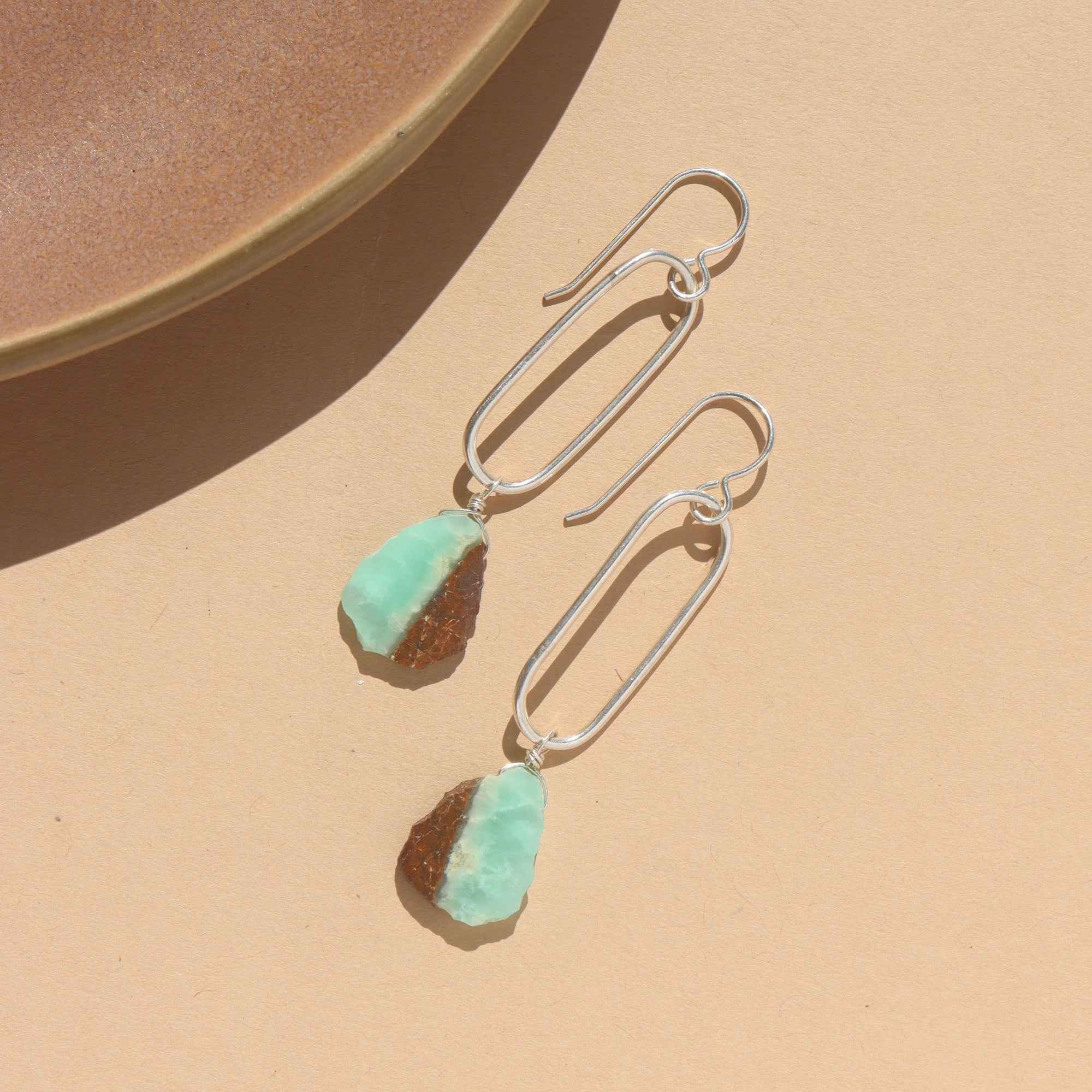 Sterling silver drop earring with a chrysophrase gemstone wire wrapped at the bottom, earring is displayed on a white dish in the sunlight. 