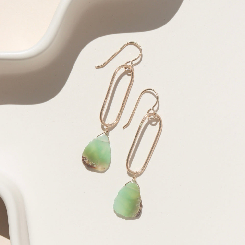 A 14k gold fill drop earring with a chrysophrase gemstone wire wrapped at the bottom, earring is displayed on a white dish in the sunlight. 