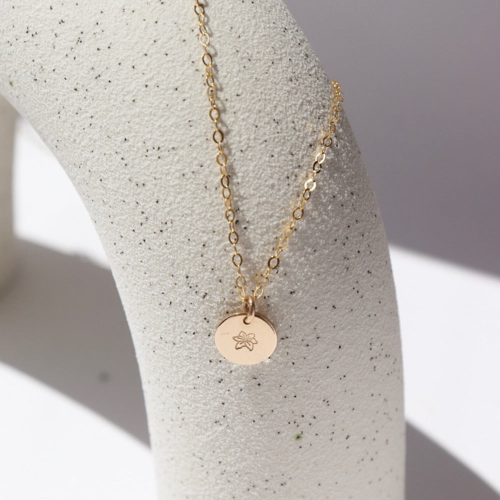 14k Gold fill Children&#39;s tiny birth flower charm necklace laid on a white jewelry stand in the sunlight.