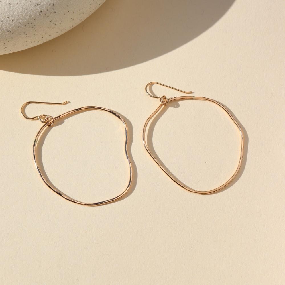 A gold large hoop earrings, each uniquely shaped, hang elegantly from simple hooks. Earrings are displayed on a cream backdrop. 