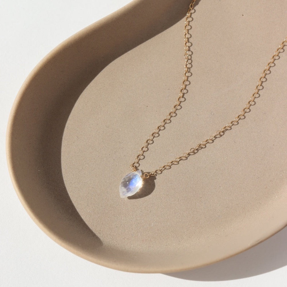 faceted marquise moonstone gemstone anchored on a solid gold chain and laying in the sunlight on a ceramic dish.