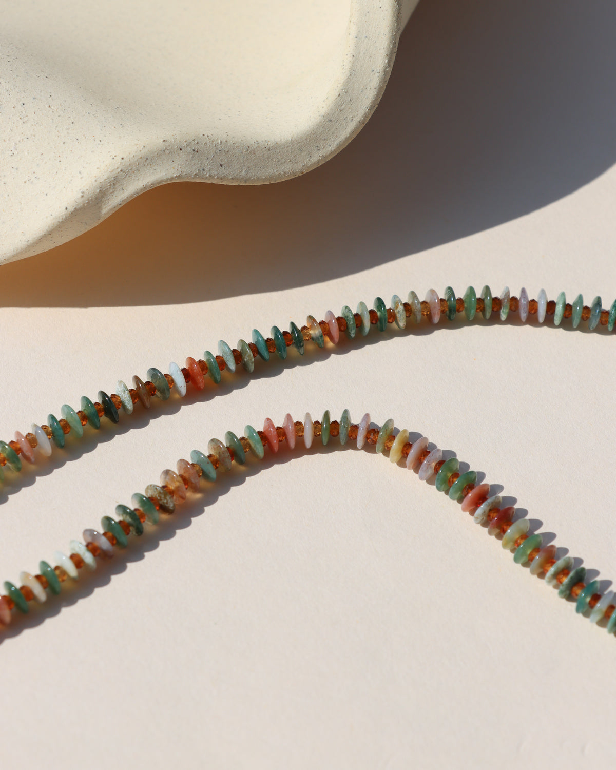 Close-up of a multicolored gemstone disc bead necklace featuring green, turquoise, blush, cream, and amber stones in warm sunlight.