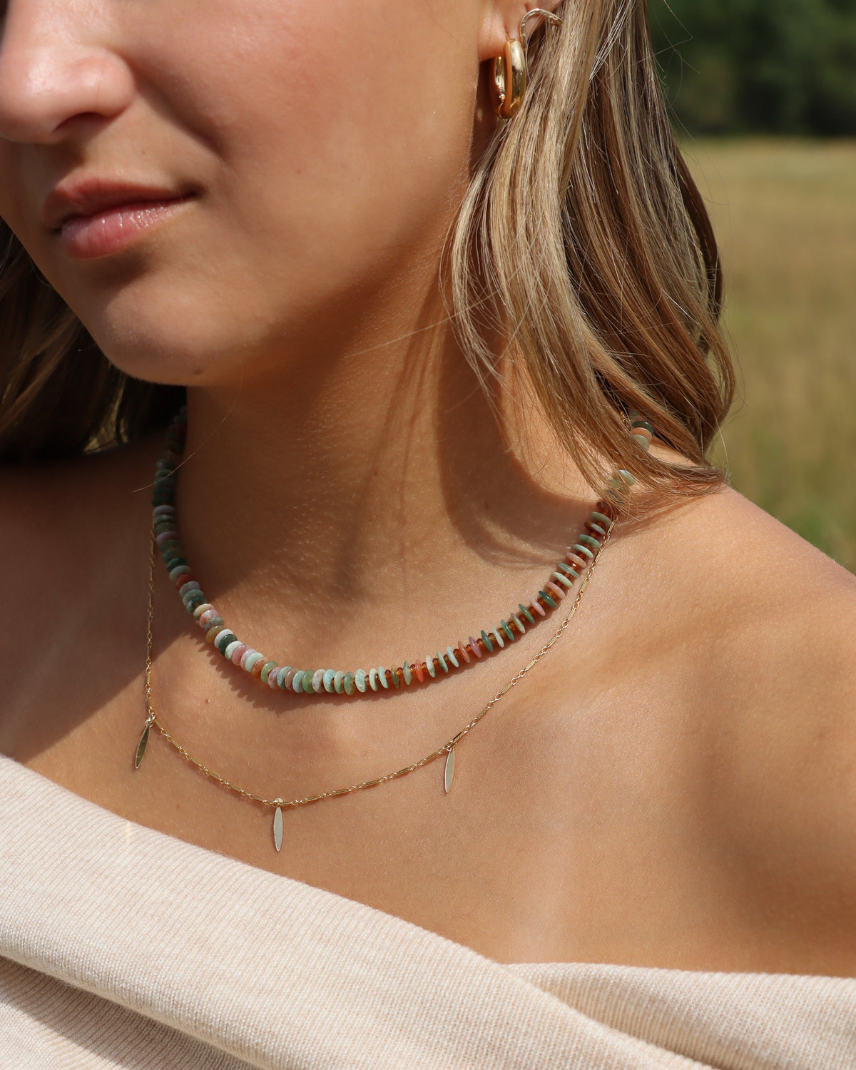 Multicolored gemstone disc bead necklace in earthy green, pink, and amber tones, layered with a delicate gold charm necklace.