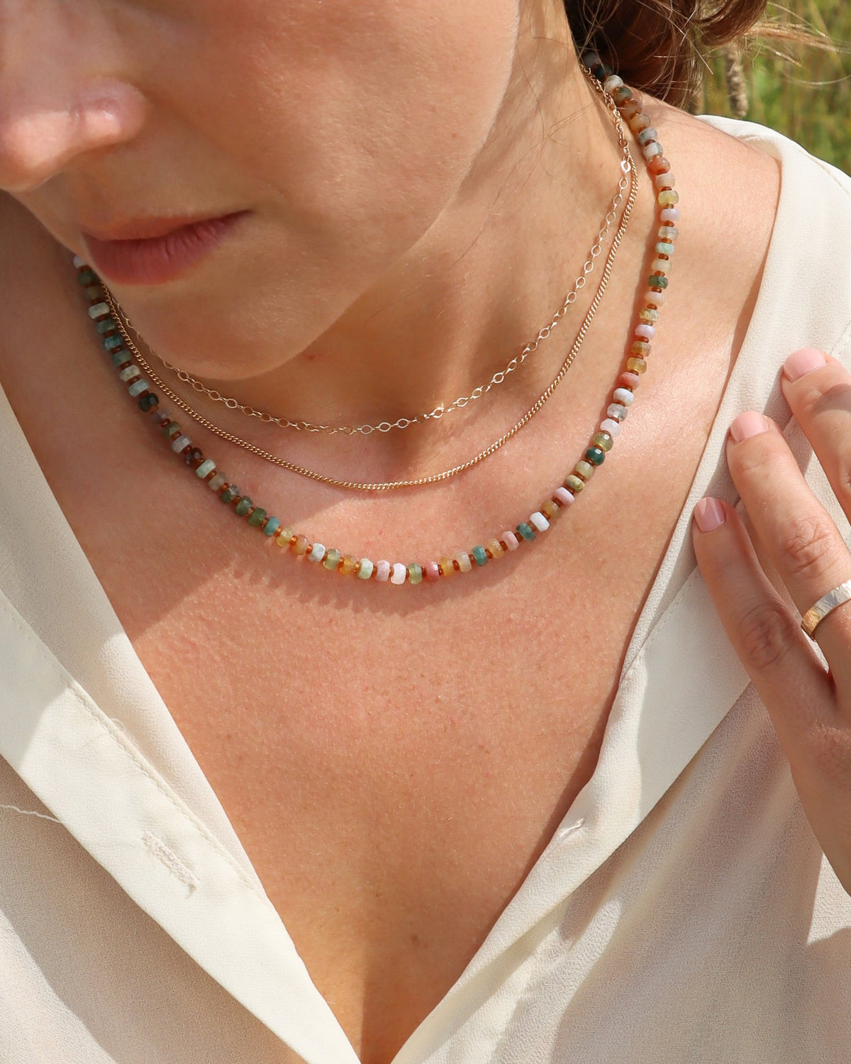 Multicolored beaded necklace in soft green, pink, cream, and earthy tones, layered with two delicate gold chains and worn with a white top.