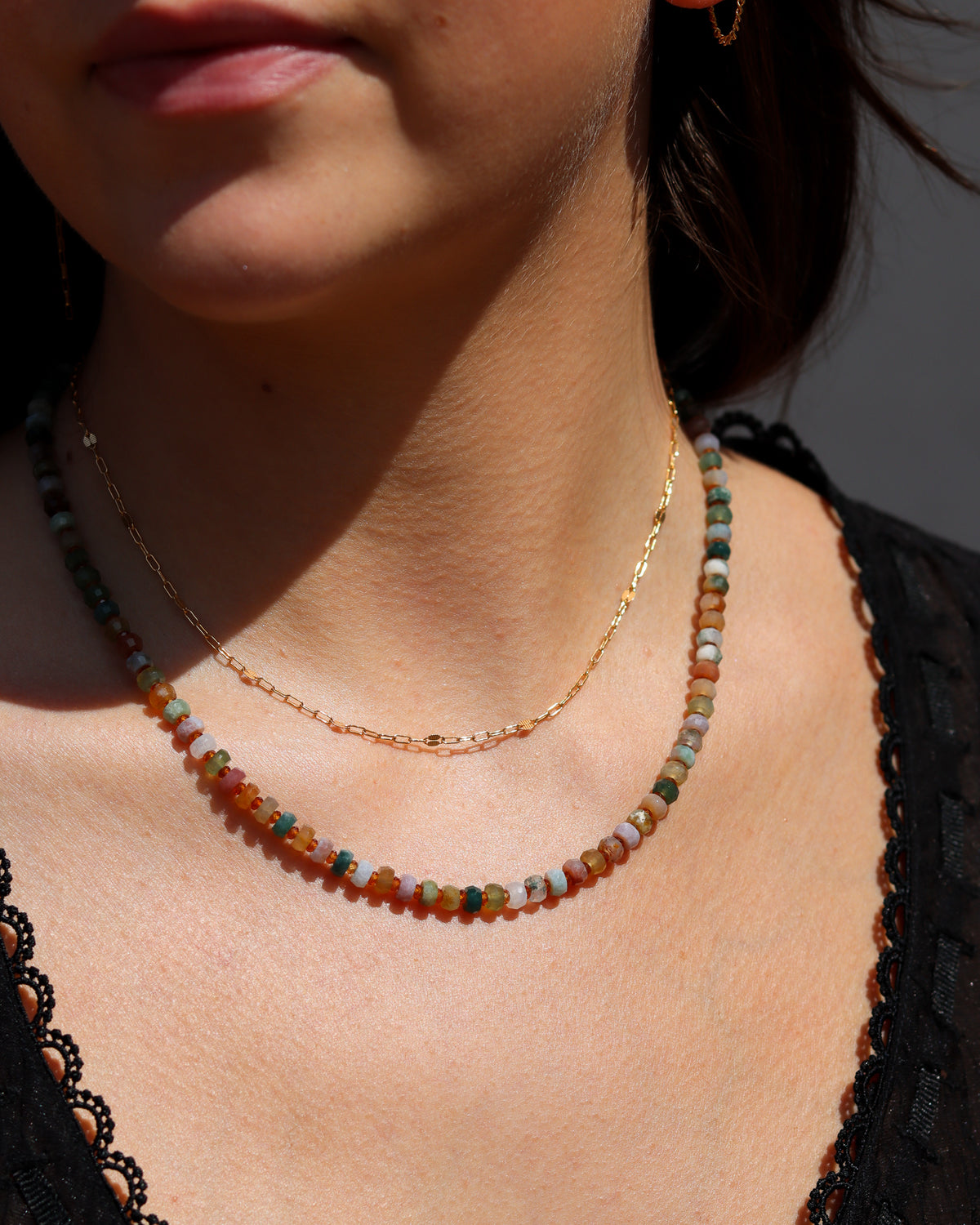 Multicolored beaded necklace in green, pink, cream, and earthy tones layered with a delicate gold chain, worn with a black top in warm sunlight.