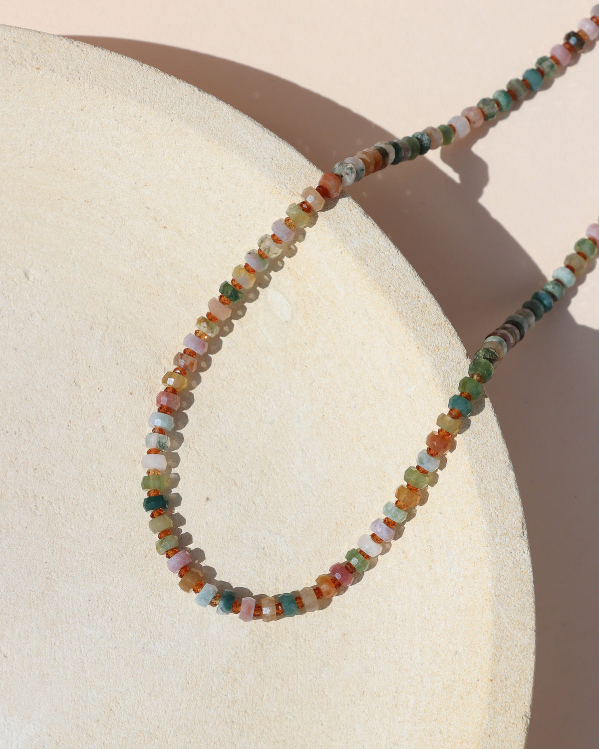Multicolored gemstone bead necklace in green, pink, cream, blue, and warm earthy tones, draped over a textured cream ceramic form in sunlight.