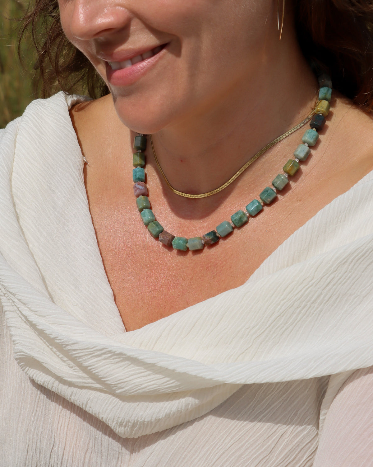 Chunky multicolored stone bead necklace in shades of turquoise, green, blue, and earthy tones, layered with a gold chain and worn with a white top.