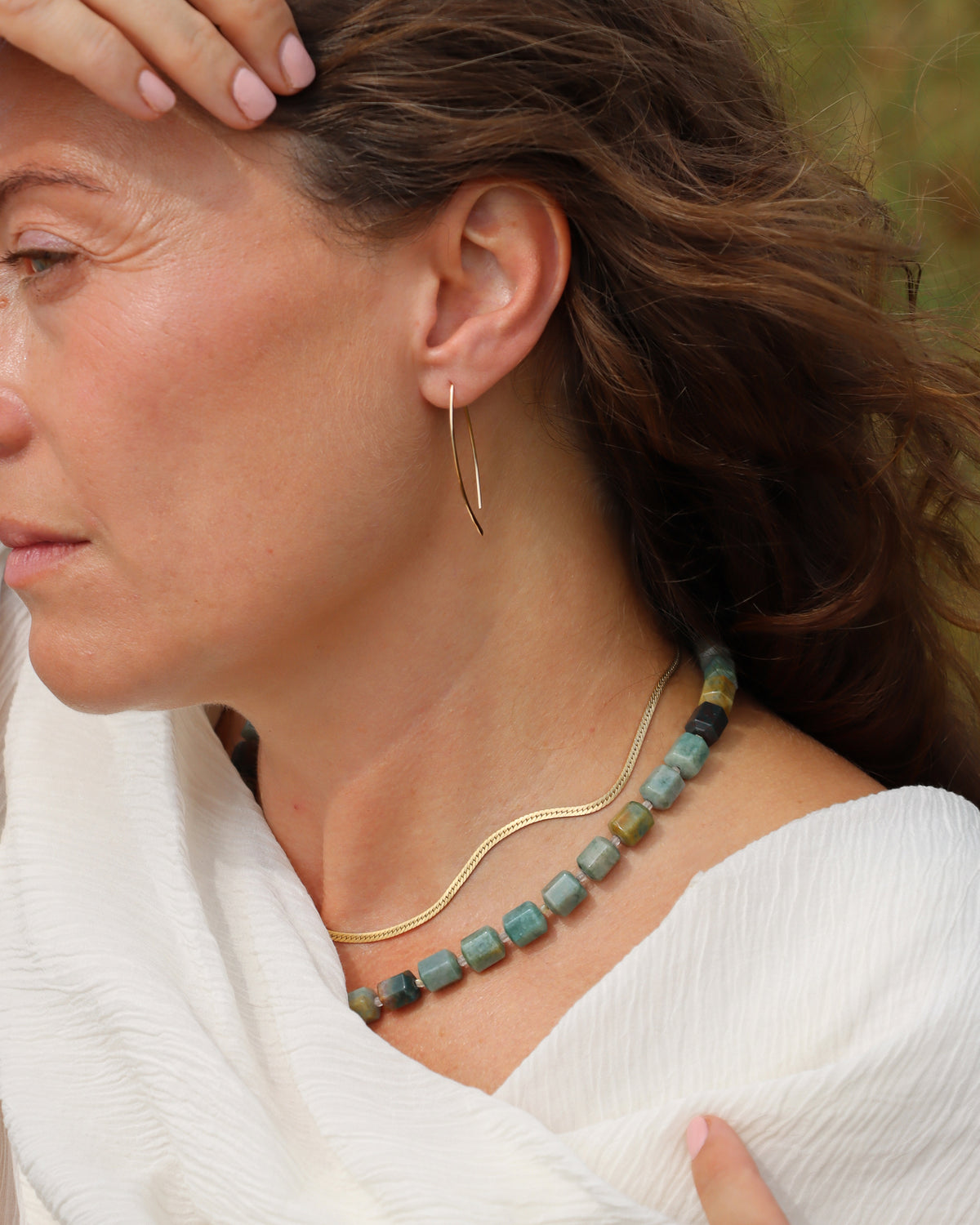Chunky multicolored stone bead necklace in turquoise, green, blue, and earthy tones, layered with a gold chain and worn outdoors.