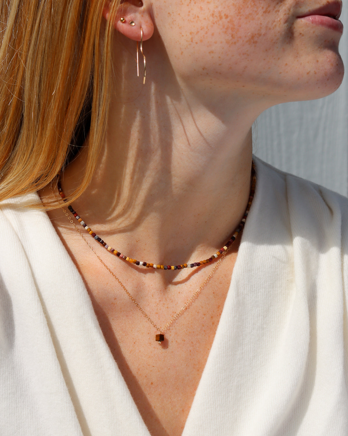 Model wearing a multicolored gemstone beaded necklace in warm earthy tones, layered with a delicate gold pendant necklace and styled with a white top.