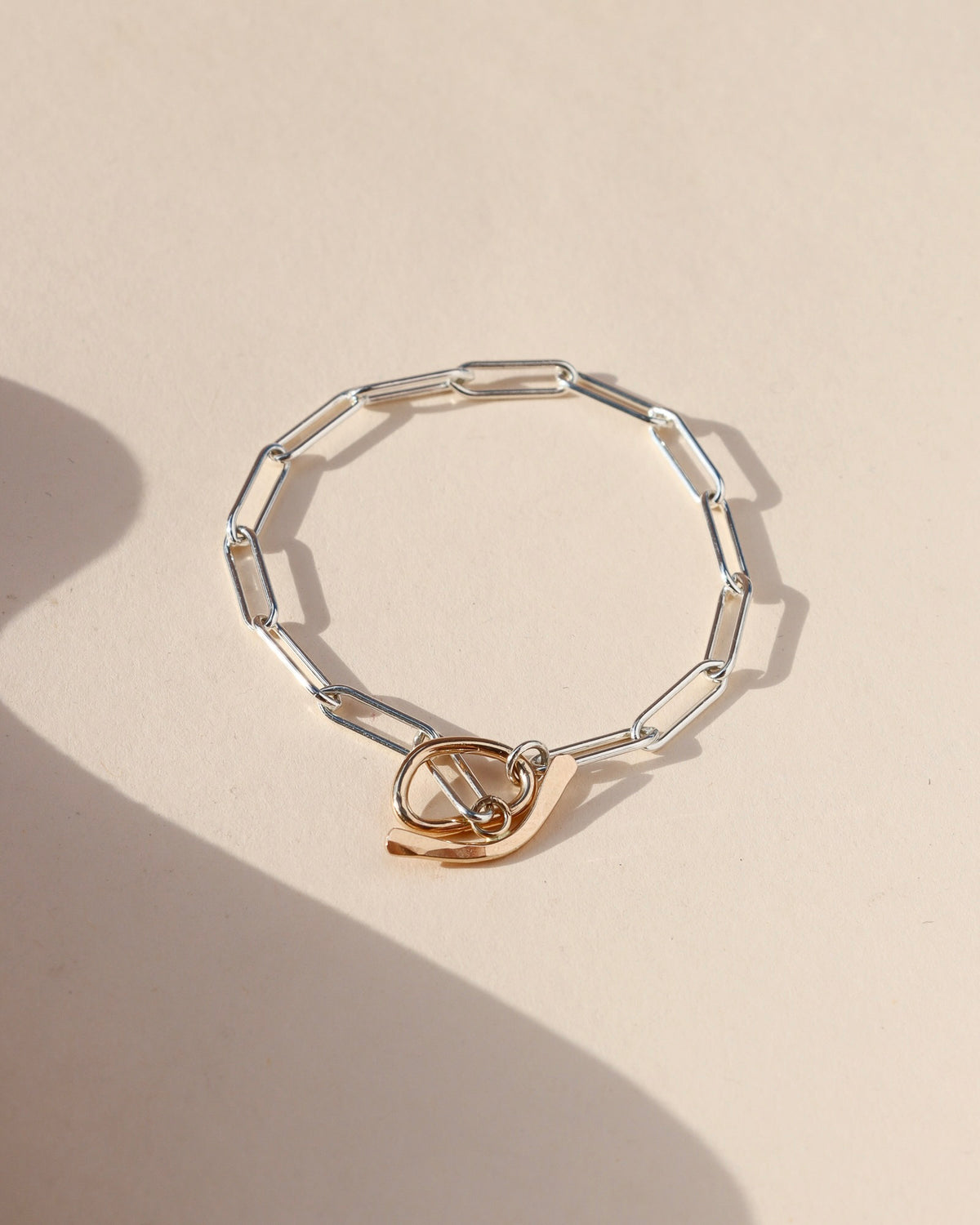 Flux Toggle Bracelet featuring elongated sterling silver paperclip chain links and a sculptural gold fill toggle clasp, displayed on a cream ceramic surface in natural sunlight.