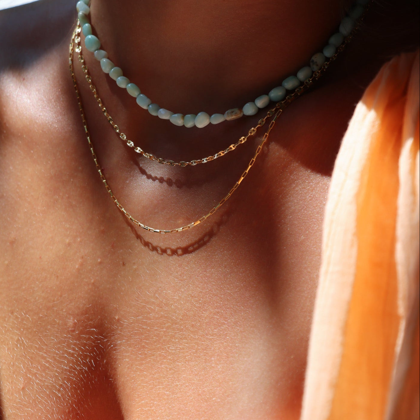 Close-up lifestyle photo of layered necklaces featuring a natural gemstone beaded choker paired with two delicate 14k gold fill chain necklaces. Photographed on a model in warm natural sunlight.