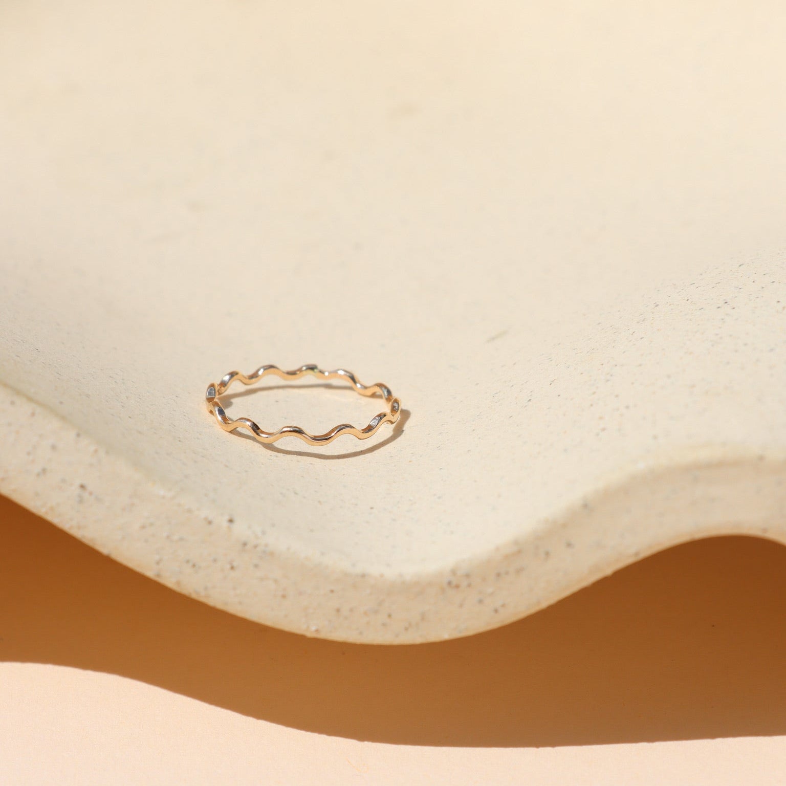 Minimalist gold ring with a gentle squiggly wave pattern, displayed inside a cream-colored textured ceramic dish with soft natural sunlight and warm shadows.