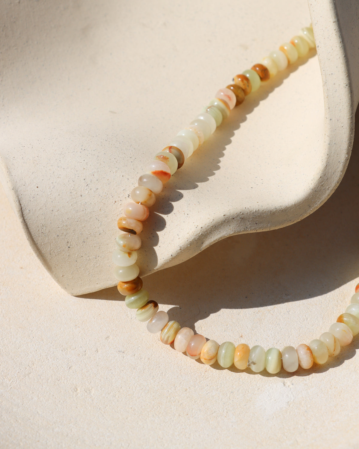 Close-up of a multicolored gemstone necklace with polished rondelle beads in pale green, ivory, peach, and caramel tones arranged against a cream textured backdrop.