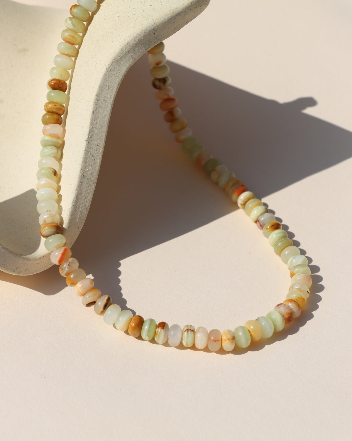 Multicolored gemstone beaded necklace featuring polished rondelle stones in soft green, cream, blush, and warm amber tones, draped over a sculptural cream display.