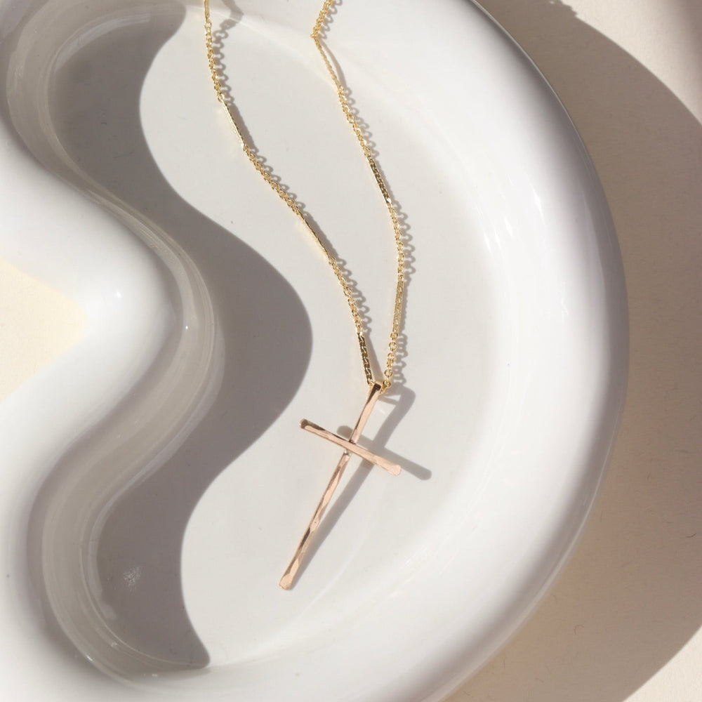 A 14k gold fill thin bar and link chain featuring a 1 1/2" hammered cross pendant. The necklace is displayed on a white dish. 