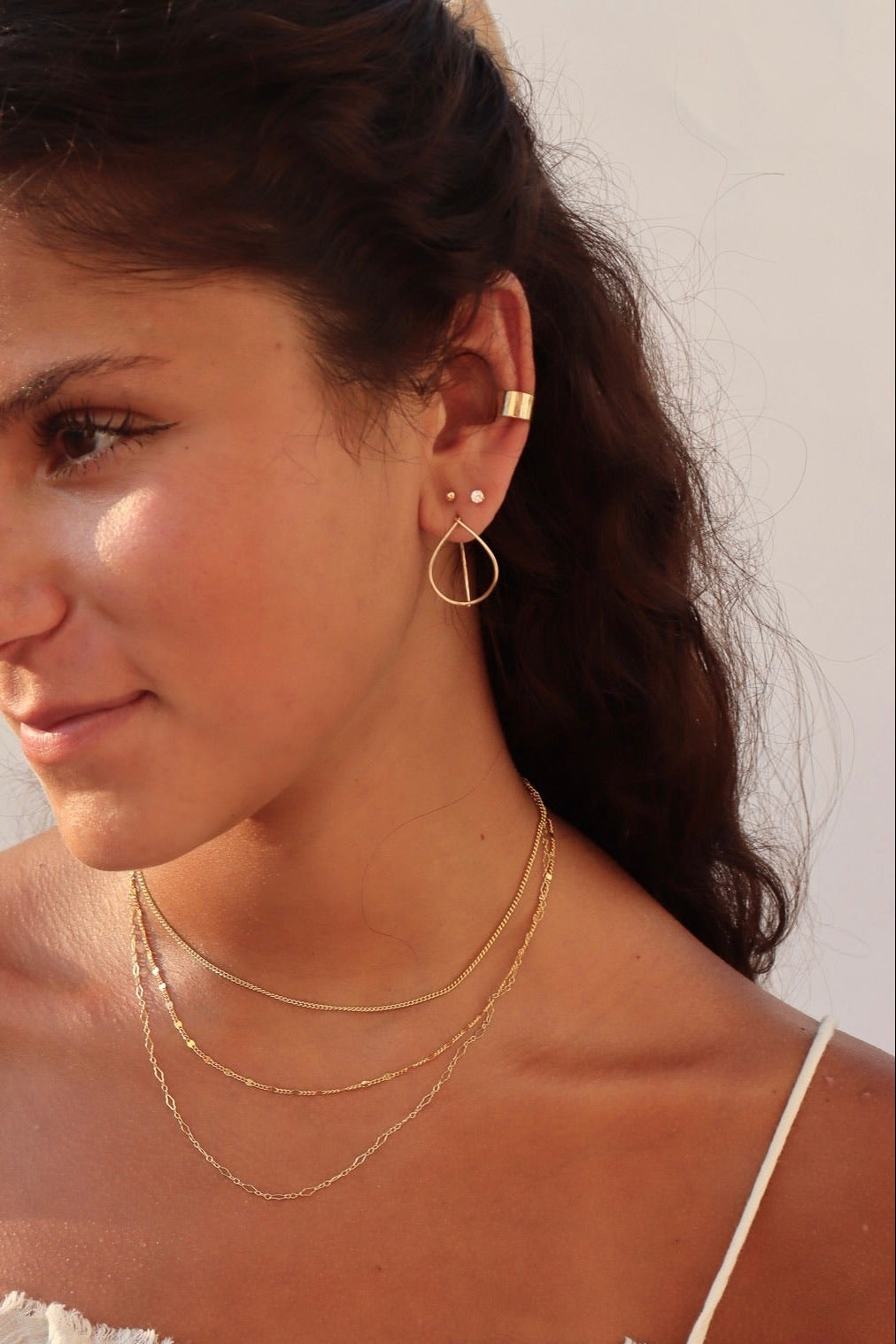 A young woman with dark hair wearing a ruffled white tank top, styling a multi-layered gold jewelry set: three delicate gold chains of varying lengths paired with geometric teardrop drop earrings, tiny stud earrings, and a gold ear cuff.