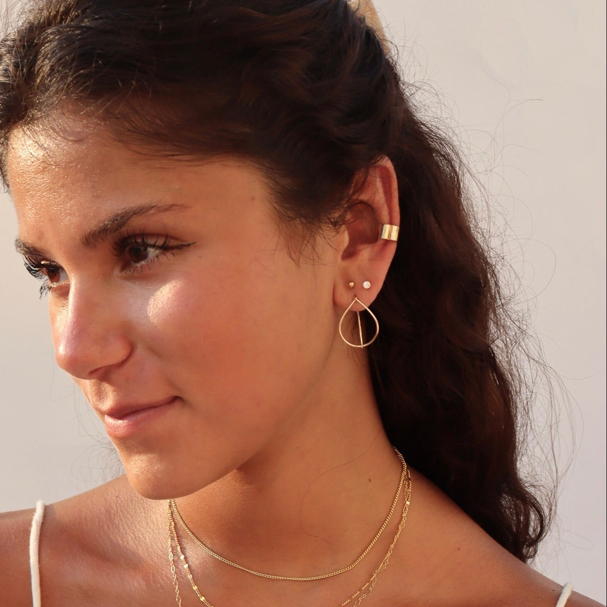 A young woman with dark hair wearing a ruffled white tank top, styling a multi-layered gold jewelry set: three delicate gold chains of varying lengths paired with geometric teardrop drop earrings, tiny stud earrings, and a gold ear cuff.