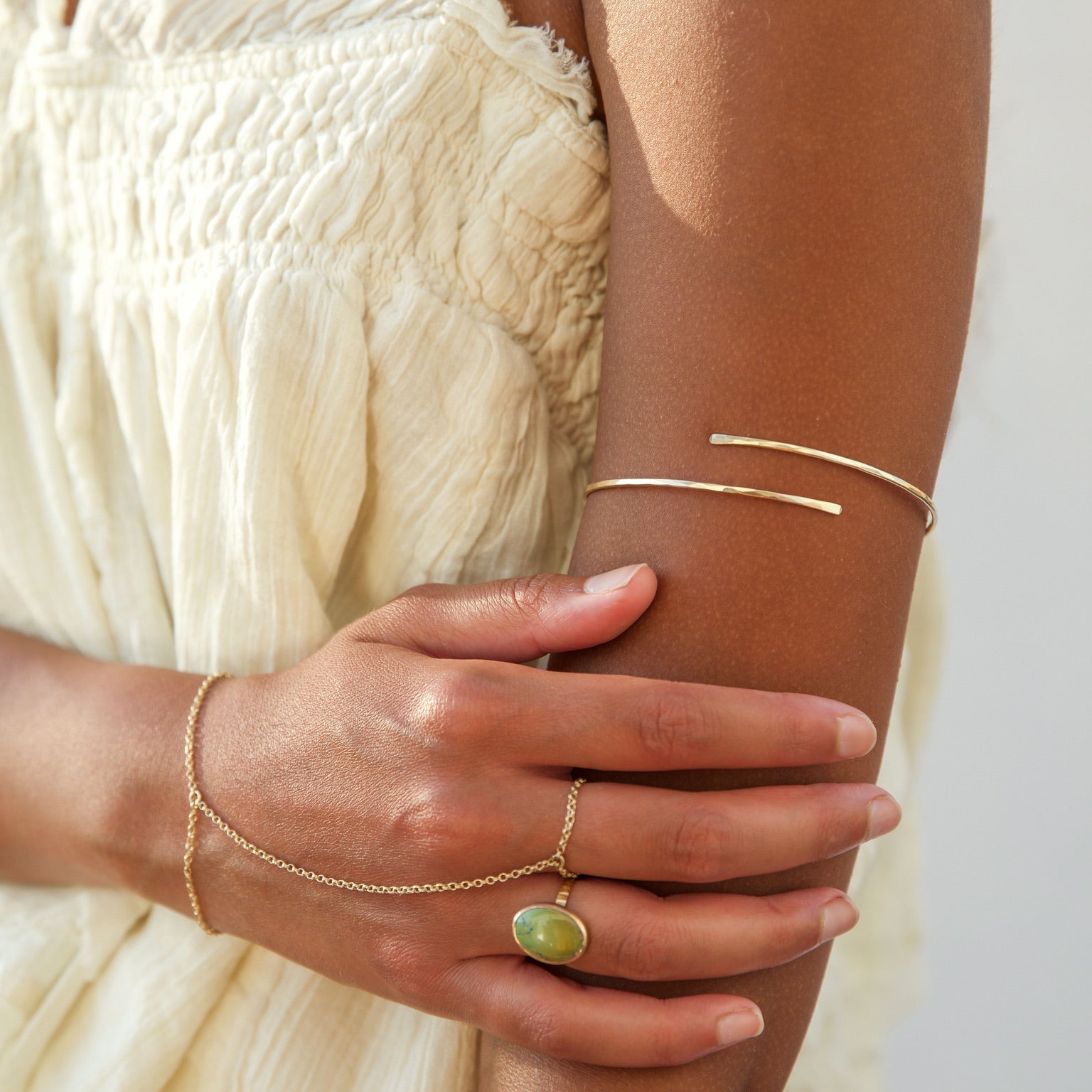 Close-up shot of a woman in a white textured dress resting her hand on her arm, styled with boho gold jewelry: a sleek open wrap upper arm cuff, a delicate gold hand chain connecting her wrist to a middle-finger ring, and an oval green gemstone ring on her index finger