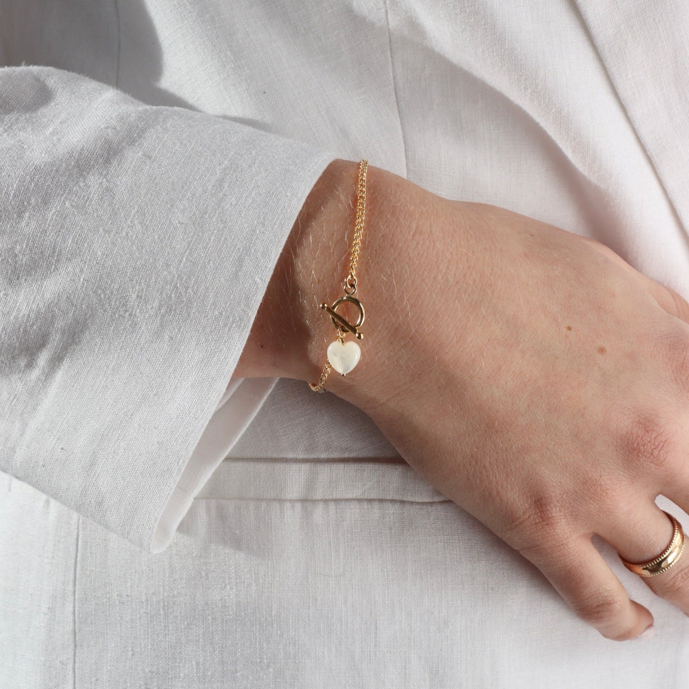 Model wearing the mother of pearl toggle bracelet. This bracelet features a gold curb chain with a toggle clasp opening with a mother of pearl heart at the end. 