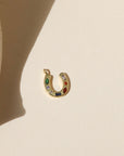 Gold horseshoe shaped charm with green, clear, blue, and red CZ gems embedded within, laid on a beige background in the natural sunlight. 