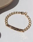 14k gold fill Alexandra Name Plate laid on a tan plate in the sunlight. This Bracelet features a simple hammered Name plate that you get to customize with any character of your choice with 8 initials. The plate is connected by the Alexandra chain.