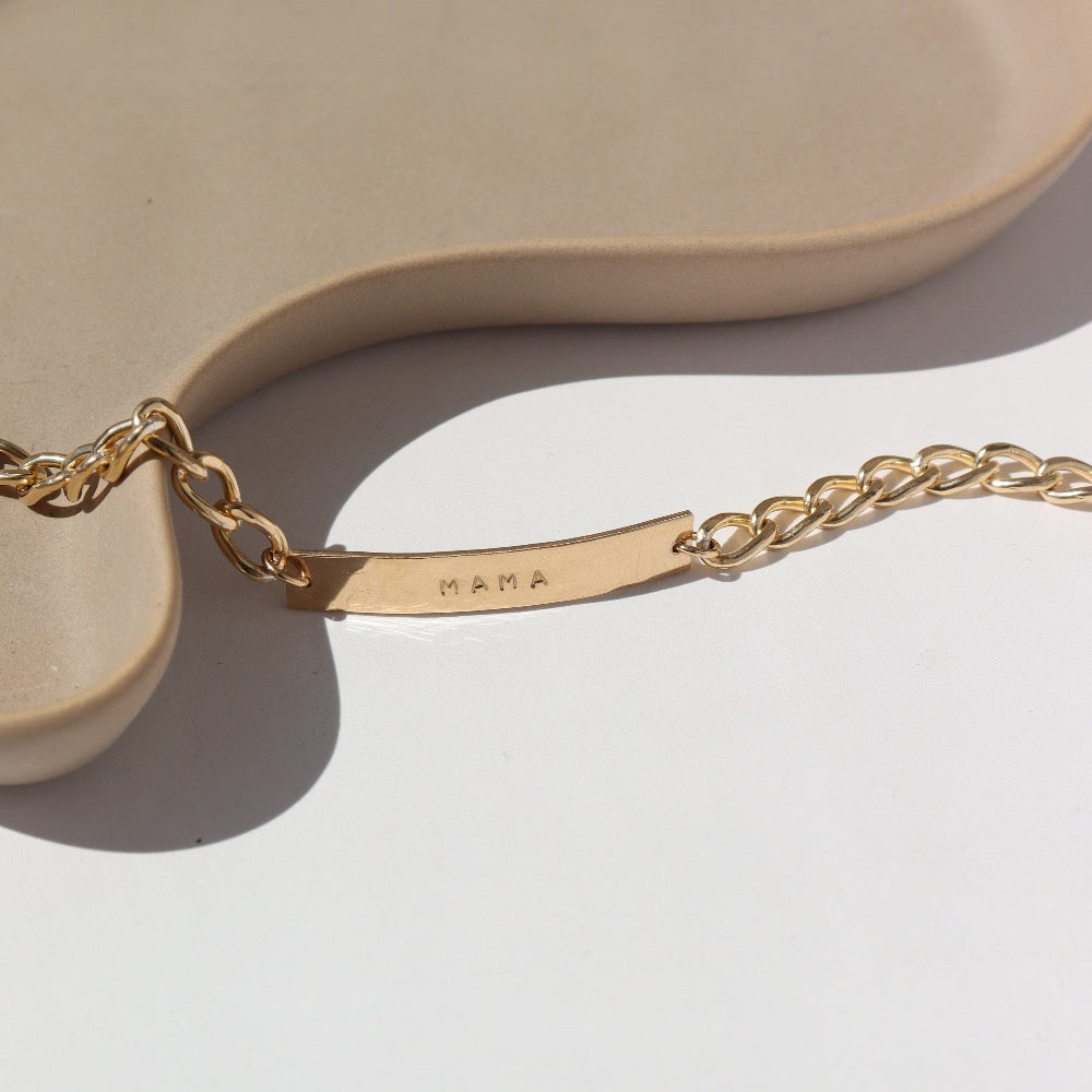 14k gold fill Alexandra Name Plate laid on a tan plate in the sunlight. This Bracelet features a simple hammered Name plate that you get to customize with any character of your choice with 8 initials. The plate is connected by the Alexandra chain.