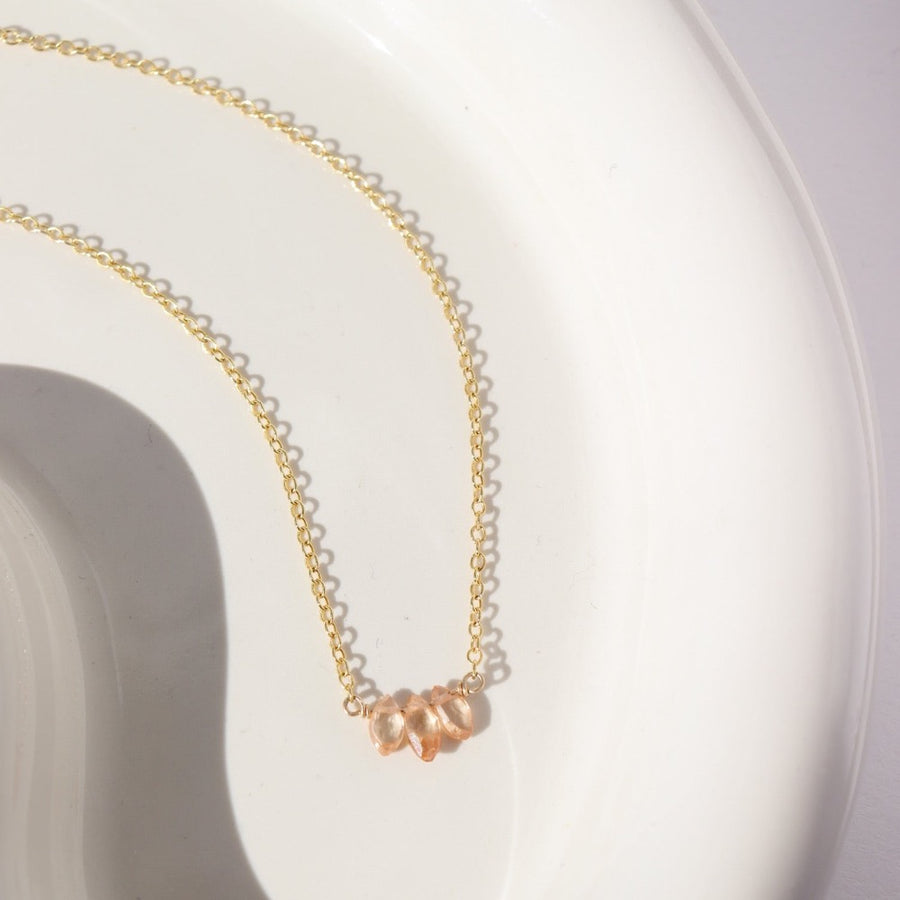 14k gold fill Gaia Necklace laid on a white plate in the sunlight. This necklace features the simple chain and is then met together by three zircon stones. 