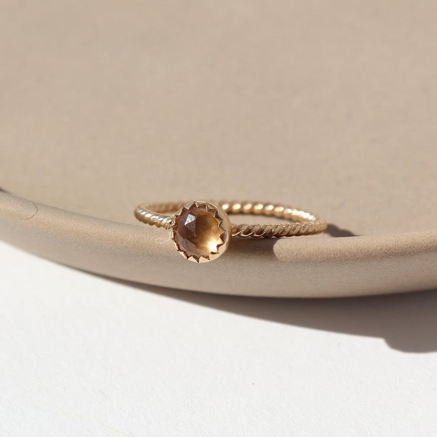 14k gold fill Gaia Ring laid on a tan plate in the sunlight. This ring features a Spiral band with a zircon gemstone. 