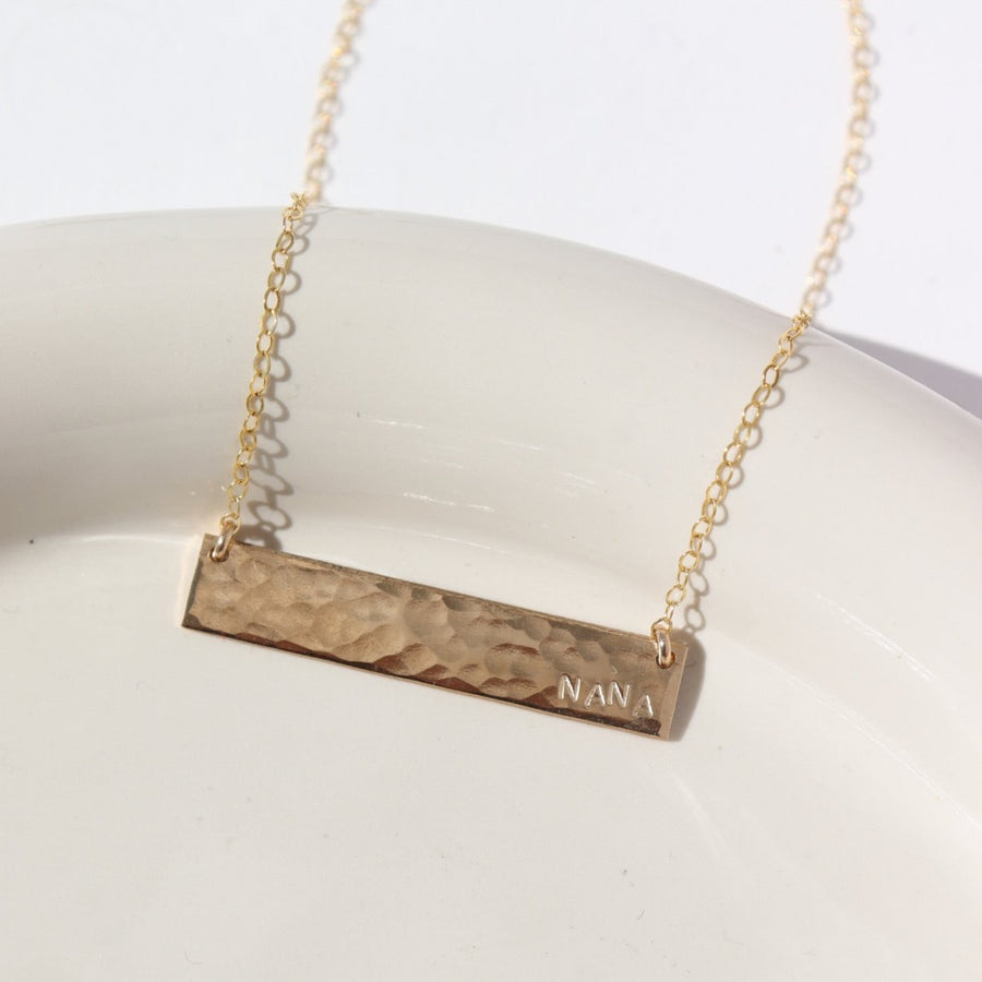 14k gold fill Matriarch Necklace laid on a tan plate in the sunlight. This necklace features either the simple chain and or the April chain and the word "NANA". you can have the option of a smoother or hammered.