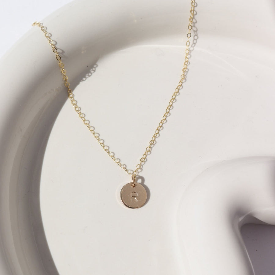 14k gold fill Tiny Monogram Disc Necklace laid on a white plate in the sunlight. This necklace features our simple chain with a disc pendent with a letter stamped on it.