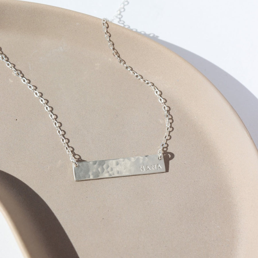 925 Sterling Silver Matriarch Necklace laid on a tan plate in the sunlight. This necklace features either the simple chain and or the April chain and the word "NANA". you can have the option of a smoother or hammered.