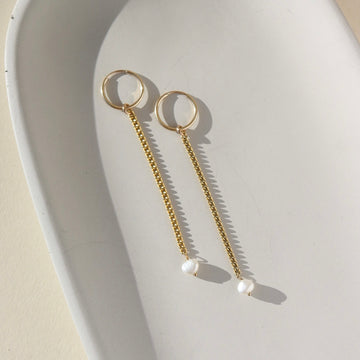 14k gold fill pearl rain drops laid on a white plate in the sunlight. This earring features our Goldie classic hoop with the La Mer chain and a tiny pearl gemstone dangling from it.