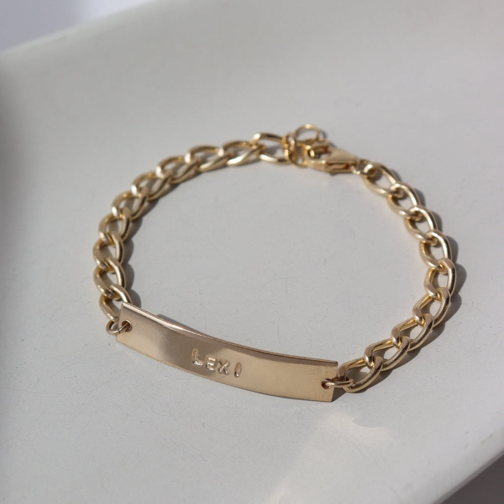 14k gold fill Alexandra Name Plate laid on a tan plate in the sunlight. This Bracelet features a simple hammered Name plate that you get to customize with any character of your choice with 8 initials. The plate is connected by the Alexandra chain.