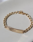14k gold fill Alexandra Name Plate laid on a tan plate in the sunlight. This Bracelet features a simple hammered Name plate that you get to customize with any character of your choice with 8 initials. The plate is connected by the Alexandra chain.
